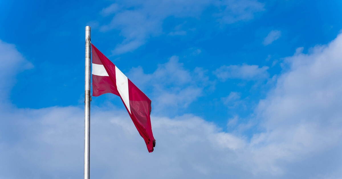 Flaga Łotwy z czerwono-biało-czerwonymi pasami powiewająca na tle błękitnego nieba z chmurami