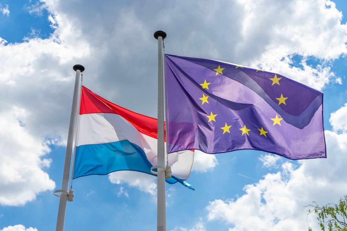 Flaga Luksemburga z czerwono-biało-niebieskimi pasami obok flagi Unii Europejskiej na tle zachmurzonego nieba
